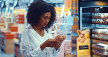 African American pleasant young woman standing by shelves with organic farmers products, choosing eggs in grocery store