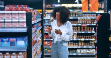 Dark-skinned pretty woman checking shopping list on smartphone, browses the web for info about food displayed for sale