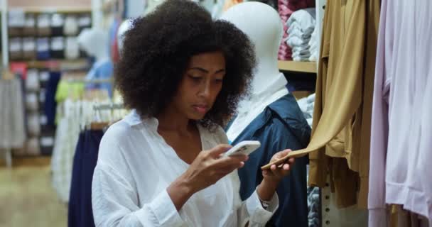 Афроамериканка Сознательный Покупатель Телефону Проверяет Информацию Текстильном Качестве Рубашки Стойке — стоковое видео