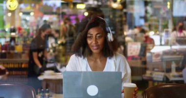 Dark-skinned pretty woman wearing white casual clothes and wireless headphones, drinks coffee from paper cup and using her laptop, browsing websites, surfing the internet, relaxing in a cozy cafeteria