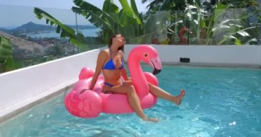 Happy female 25 in blue bikini and red sunglasses relax at vacation travel, swimming in pool on trendy inflatable pink flamingo. Smiling female traveler enjoying luxury relax summer holidays sunny day