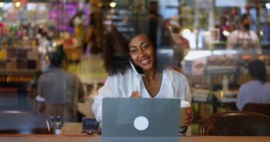 Busy overworked multi-ethnic young woman, freelancer in casual wear, talking on mobile phone and telecommuting on laptop, enjoying her coffee break in cozy cafeteria. Businesswoman. Entrepreneur.