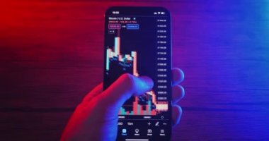 Close up hands of crypto trader holding mobile phone analyzes cryptocurrency price. Crypto investor makes money using mobile phone speculating on cryptocurrency, at night in neon light. Mobile trading