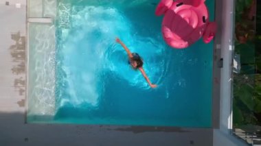 A young woman having fun, relax in pool, spins and splashes with water on vacation at summer resort. Drone aerial shot woman actively relax small swimming pool hotel at traveling vacation. Top view