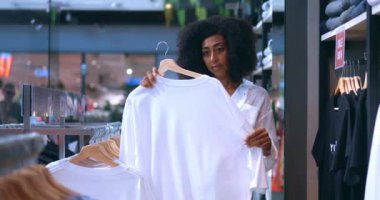 Happy young African American woman customer with shaggy hairstyle, in casual wear, holding stylish white t-shirt, smiling while enjoying shopping on her free time, in a modern shopping mall
