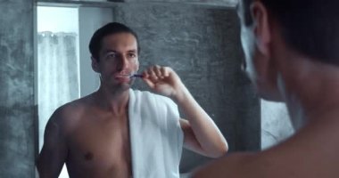 Young man brushing teeth in the bathroom. Man with towel on shoulder looks in the mirror and brushing his teeth in the morning after waking up. Dental care concept. Head shot mirror reflection