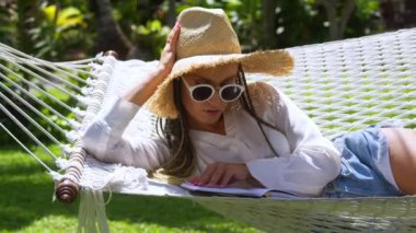 Hasır şapkalı kadın hamak dinleniyor ve yaz tatillerinde kitap okuyor. Tropikal bir ülkede yaz tatilinde dinlenen kadın turist, tropik tatil beldesindeki hamakta dinlenen kitap okuyor..