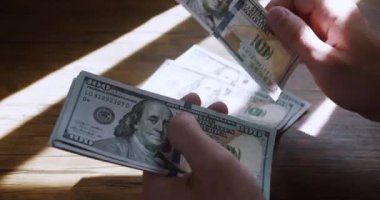 Güneşli bir günde maaş gününde ahşap masadaki 100 dolarlık banknotları saymak. İş adamı elleri kağıt para sayıyor. Erkek, 100 dolarlık banknotları sayarak eğleniyor. Para kazanıyorum.