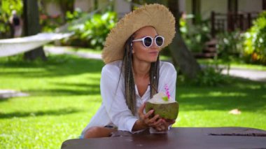 Elinde hindistan cevizi olan hasır şapkalı ve güneş gözlüklü kadın turist, tropik tatil beldesinde yaz haftasonunun tadını çıkarıyor. Ellerinde tropikal içeceklerle rahat bir kadın tropikal tatil köyünde yaz haftasonunun tadını çıkarıyor..