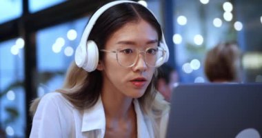 Young Asian Business Project Manager in Headphones Working on Laptop Computer in Busy Modern Office. Beautiful Diverse Multiethnic Female Specialist Concentrated Writing Business Strategy at Laptop.