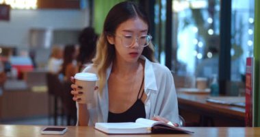 Genç Asyalı 20 kadın modern kafede rahatlıyor, kitap okuyor ve lezzetli kahve içiyor. Gözlüklü güzel Asyalı kadın kahve dükkanında kitap okuyor ve latte içiyor. Kitap okuma kavramı.