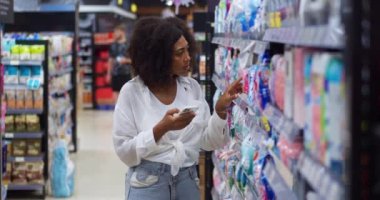 Conscious housewife using mobile app on phone, browses on web for information about washing powders displayed for sale