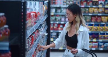 Young woman 20 teenager in glasses in grocery supermarket chooses groceries and consumer goods. Asian woman consumer with shopping cart in hand buys grocery in food supermarket. Shopping at food store