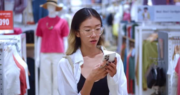 戴眼镜的亚洲女人在室内服装店里滚动智能手机 用智能手机在成排成排的服装店之间穿行的亚洲女性形象 同时在网上购买价格更低的商品 — 图库视频影像