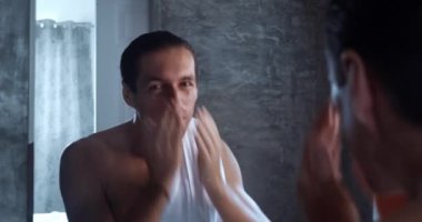 Smiling man washes and wipes himself with a towel while standing in the bathroom in the morning in front of a mirror. Mirror reflection young happy bare man washing and wiping face with towel.