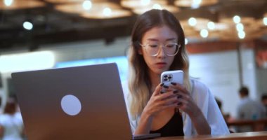 Asian young female student use mobile phone app and computer to work or study, sitting in coffee shop. Teenager woman in glasses looking at mobile phone screen and use laptop computer in stylish cafe.