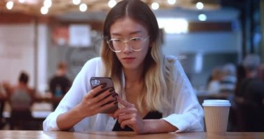 Beautiful young asian woman 20 using smartphone while sitting at cafe. Asian female student in eyeglasses using app on mobilephone at coffee shop. Beautiful girl relaxing, chatting on mobile phone.