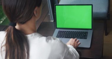 Serbest çalışan kadın tasarımcı pencerenin önündeki rahat kafede yeşil ekran maketli dizüstü bilgisayar kullanıyor. Omuzlarının arkasından görünen dişi, iç dekorasyonunda yeşil ekran krom anahtarı olan bir bilgisayar kullanıyor..