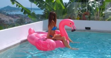 Havuzda şişirilebilir pembe flamingo şilte ile bikini içinde dinlenen güzel bir genç kadın. Lüks deniz manzaralı villa gezisinde güneşlenen mutlu kadın tropikal bir ülkede tatil yapıyor..