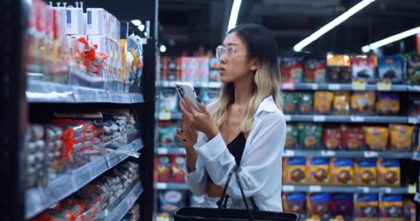 亚洲女性购物者使用手机在网上阅读有关食品的评论 计算卡路里 带着食品在杂货店购物 亚洲妇女购物者在食品超级市场选择食物 — 图库视频影像