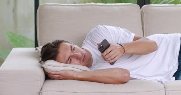 悲しい男はソファに横たわって スマートフォンにメッセージを読み取り 悪いニュースに苦しんでいます 30歳の男が家のソファに横になるのが悲しい うつ病の概念 — ストック動画