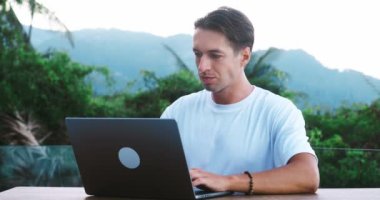 Genç erkek serbest çalışan doğa arka planında dizüstü bilgisayarda çalışıyor. Millennial erkek serbest çalışan, tropik bir cennet ülkesinden dizüstü bilgisayarın başında çalışıyor. Uzak çalışma kavramı.