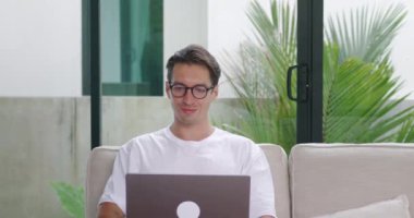 Gözlüklü erkek milenyum öğrencisi evinde kanepede oturur ve online çalışmak için dizüstü bilgisayar kullanır. Erkek öğrenci evden bilgisayarda çalışıyor. Kanepede oturuyor.