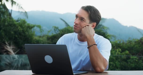 風光明媚な自然の中でノートパソコンとテーブルに座っている間 受動的な笑顔の男は新しいビジネスアイデアを夢見ています オンラインでリモートで作業することによって 事業を開始するための新しい革新的なアイデアを生成します — ストック動画