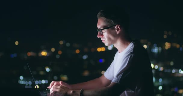 在夜市背景下 一个戴着眼镜在笔记本电脑上工作的侧视自由职业者 自由职业者在笔记本电脑上远程工作 在键盘上打字 坐在屋顶上背景音乐 — 图库视频影像