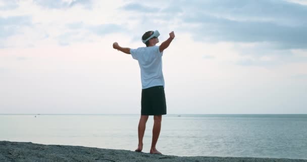 仮想世界での勝利の喜びのためにジャンプする男の勝者 仮想現実のVrメガネを使用して再生 海の背景に屋外で 日没時にビーチに旅行中のVrヘッドセットを使用して男 — ストック動画