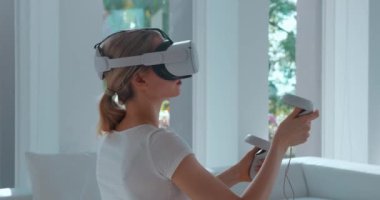 VR gözlüklü bir kadın evdeki kanepede oturup video oyunu oynamaktan hoşlanıyor. Çekici bayan oyuncu evde artırılmış realite oyunları oynamak için sanal gerçeklik kulaklığı ve kumanda kullanıyor..