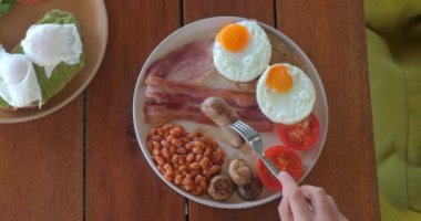 Sabah İngiliz kahvaltısı yerken, çatallı sosis yiyen bir kadın. Geleneksel İngiliz kahvaltısı sosisli, sahanda yumurta Benedict, domates, mantar, fasulye.