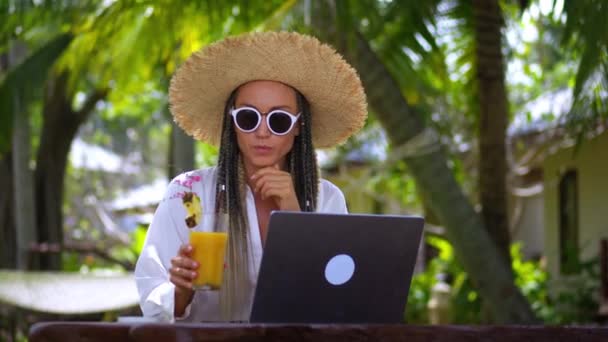 女性観光客のフリーランスは 夏の休暇に熱帯リゾートでラップトップでリモートで働いています 帽子とサングラスで女性のフリーランサーを笑顔熱帯の国でラップトップを使用して働いています リモートワーク — ストック動画