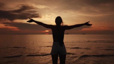 Sırtı kameraya dönük, denizde gün batımını izleyen genç bir kadının silueti. Kadın yavaşça ellerini kaldırır ve deniz günbatımının tadını çıkarırken yanlarına sertçe yayar.