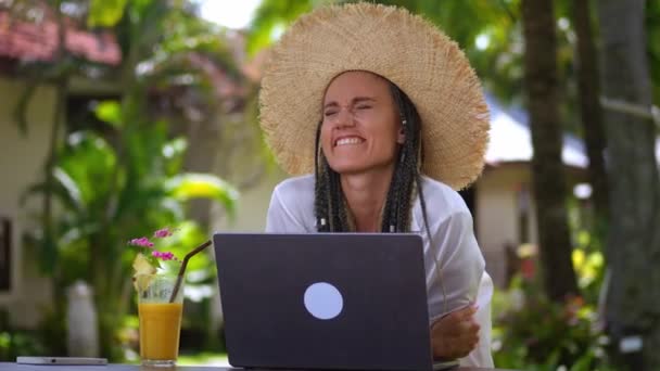 ノートパソコンの画面を見ながらわら帽子の興奮した女性が勝つことを喜びます ラップトップを見て幸せな女性観光客 夏休み中に勝利とビジネスの成功を祝うトロピカルカクテルを飲む — ストック動画