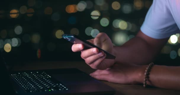 夜の街を背景にノートパソコンで仕事をしながら携帯電話にメッセージを書いているビジネスマンのクローズアップ 男性実業家は 夕方にコンピュータで作業しながら携帯電話を使用します — ストック動画