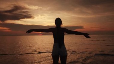 Turuncu gün batımında genç bir dişinin silueti, havada el kaldırması, denizde neşeli bir günbatımı dönüşü. Mutlu kadın seyahat ederken güzel turuncu deniz manzaralı günbatımının tadını çıkarıyor. Yaz tropik tatili