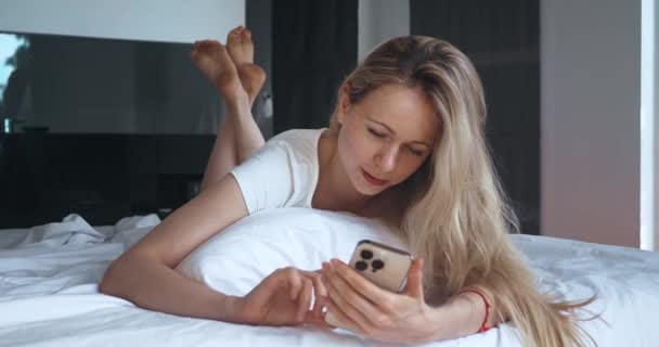 Pokojowa Milenijna Kobieta Trzymająca Smartfona Smsy Leżąc Przytulnym Łóżku Sypialni — Wideo stockowe