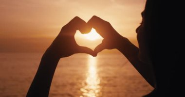 Altın gün batımında kadın eli yaklaştı. Kalbi kalp şeklinde yaptı. Gün batımında deniz üzerinde güneşi çerçeveledi. Dişi kalp işaretleri yapar. Yaz tatilinde, akşam güneşlerinde denizin tadını çıkarır..