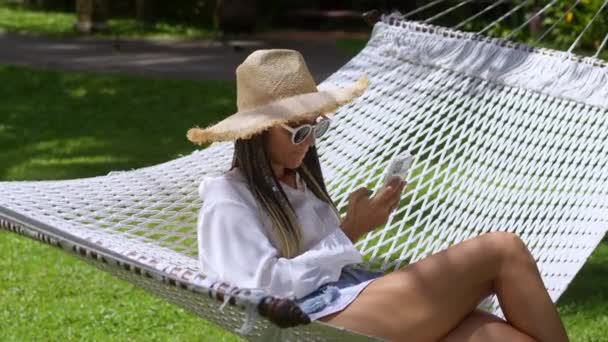 藁帽子とサングラスの女性旅行者はハンモックと夏のリゾートにあり 携帯電話を手に持っています 夏の旅行中にハンモックで休んで携帯電話アプリを使用して楽しむ女性旅行者 — ストック動画
