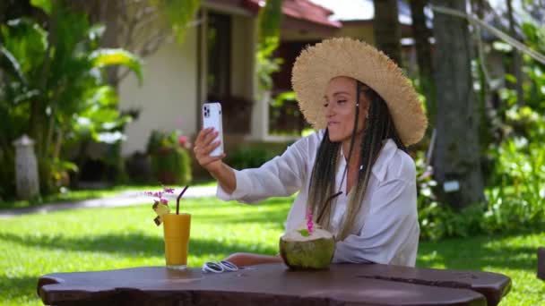 友人にスマートフォンでビデオ通話をするわら帽子の女性観光客を休憩すると 夏休みの印象が共有されます 笑顔の女性観光客が手にスマートフォンを保持します ビデオ通話 — ストック動画