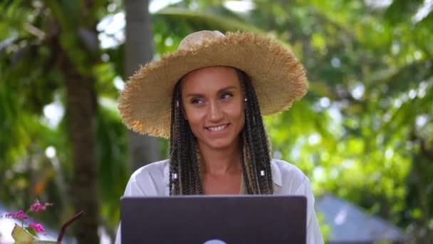 熱帯の島で夏の旅行を楽しみながら ラップトップを使用してわら帽子で女性旅行者を笑顔 熱帯の屋外カフェでノートパソコンをスクロールすることを楽しむ女性旅行者 デジタル遊牧民の概念 — ストック動画