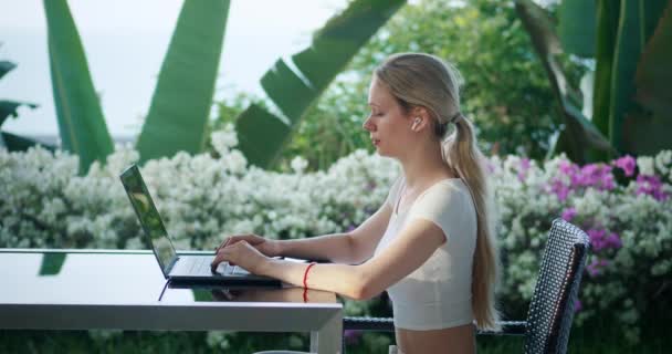 年轻的商业女性数码游牧民族坐在户外咖啡馆里 在笔记本电脑前工作 在笔记本电脑上工作的白种人女 戴着耳机 一边旅行一边远程做自由职业工作 — 图库视频影像