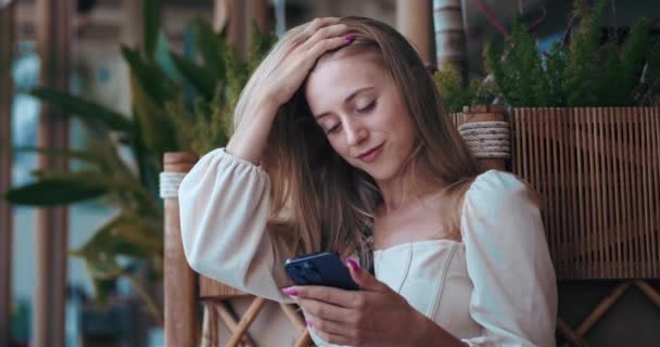 笑顔の若い女性は 通りのカフェでベンチに座って モバイルアプリケーションを使用して 彼女のボーイフレンドとスマートフォン 浮気やチャットを使用しています 携帯電話のチャットを使用して手でスマートフォンと幸せなブロンドの女性オンライン — ストック動画