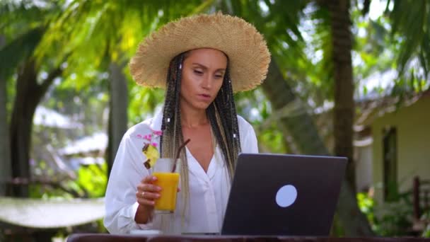 デジタルノマドとしてラップトップでオンラインで働くストロー帽子の女性旅行者は 夏休み中にコンピュータ上のリモートワークを楽しんでいる間 マンゴーシェイクを飲んでいます オンラインビジネス デジタルノマドコンセプト — ストック動画