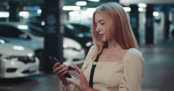 女性は 最新のニュースの使用モバイルアプリケーションに囲まれた駐車場に立っています スクリーンの柔らかい光は彼女の表面の失われた反射を投げます 彼女は使用モバイル アプリケーションに焦点を当てています — ストック動画