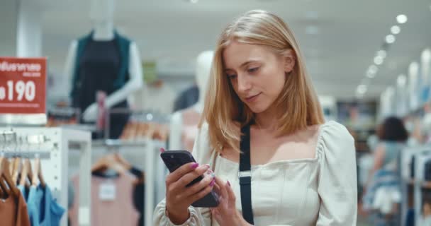 ショッピングコンセプト 女の子は衣料品部門のスマートフォンを 飛ばすことに集中してる 彼女は彼女のガジェットの買い物リストをチェックする スーパーマーケットに立っている女性は 支出を追跡します — ストック動画