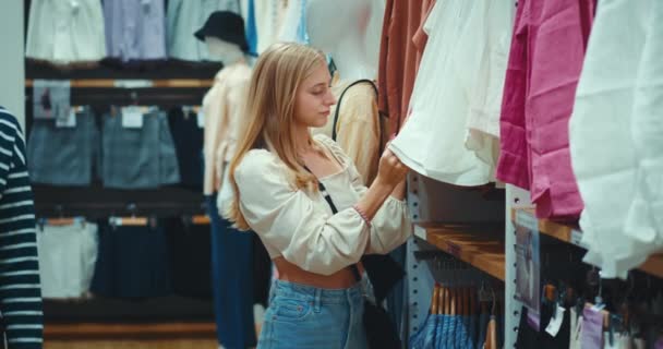 多愁善感的女性会检查商店衣架上的有机棉衬衫 女性站在商场有机棉布的服装店时 触摸面料并检查其触摸程度 — 图库视频影像