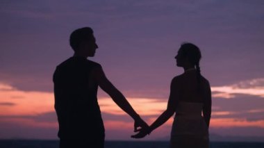 Silüet romantik çift, bir erkek bir kadını gün batımının arka planına doğru elinden tutar. Günbatımında romantik bir çift akşam denizin arka planına karşı. Romantik randevu kavramı.
