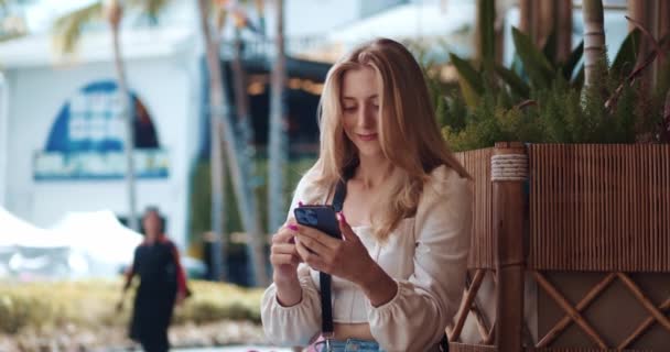 ポジティブな若い女性学生は 屋外のストリートカフェでベンチで休んでいる間 スマートフォンを使用して モバイル画面を見ます スマートフォンで笑顔の女性学生 キャンパスでベンチに座って — ストック動画